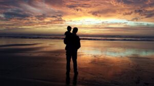 A father holds his son in silhouette on a beach watching a setting sun. 
