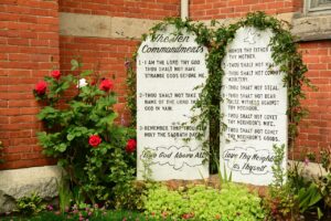 Let Churches Teach Commandments blog post by Ken Walker Writer. Pictured: The Ten Commandments posted outtsidee a brrick building, with greenery growing over them.