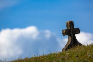 Eternal Promise Amid Tragedy blog post by Ken Walker Writer. Pictured: A cross tombstone sitting on a hill of green grass with a blue sky in the background.