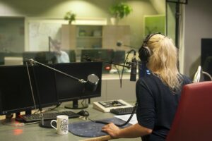 Pictured: A woman sitting at a microphone, her back to the camera, in a booth at a modern radio station.