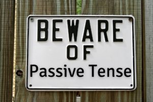 Getting Active in Your Writing blog post by Ken Walker Writer. Pictured" A white and black sign on a wooden fence that reads Beware of Passive Tense