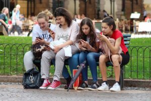 Loneliness and the Need for Personal Connections blog post by Ken Walker Writer. Pictured: Four women of various ages sitting on a park bench on a nice day, scrolling on their phones.