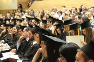 Pictured: Graduates in black cap and gown sitting in an auditorium.