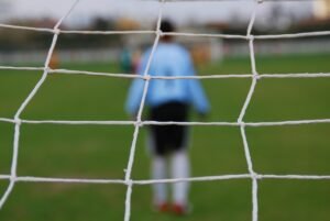 The Ascension of Soccer Fever blog post by Ken Walker Writer. Pictured: Seen through the back of a soccer goal net a goalie stand ready.