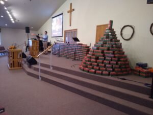 A Shoebox That Changed a Life blog post by Ken Walker Writer. Pictured: A church stage with a pyramid and other piles of shoeboxes for Operation Christmas Child. A cross can be seen on the wall behind them.