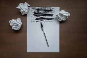 A Novel Idea blog post by Ken Walker Writer. Pictured: A piece of paper and a pen sitting on a desk with crumbled paper surrounding them. On the paper words have been scribbled out.