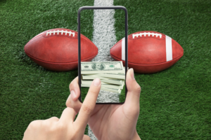 Gambling Ruining Sports blog post by Ken Walker Writer. Pictured: Two American footballs sit on a field. A pair of hands holds a phone with money on the screen as if betting on the game.
